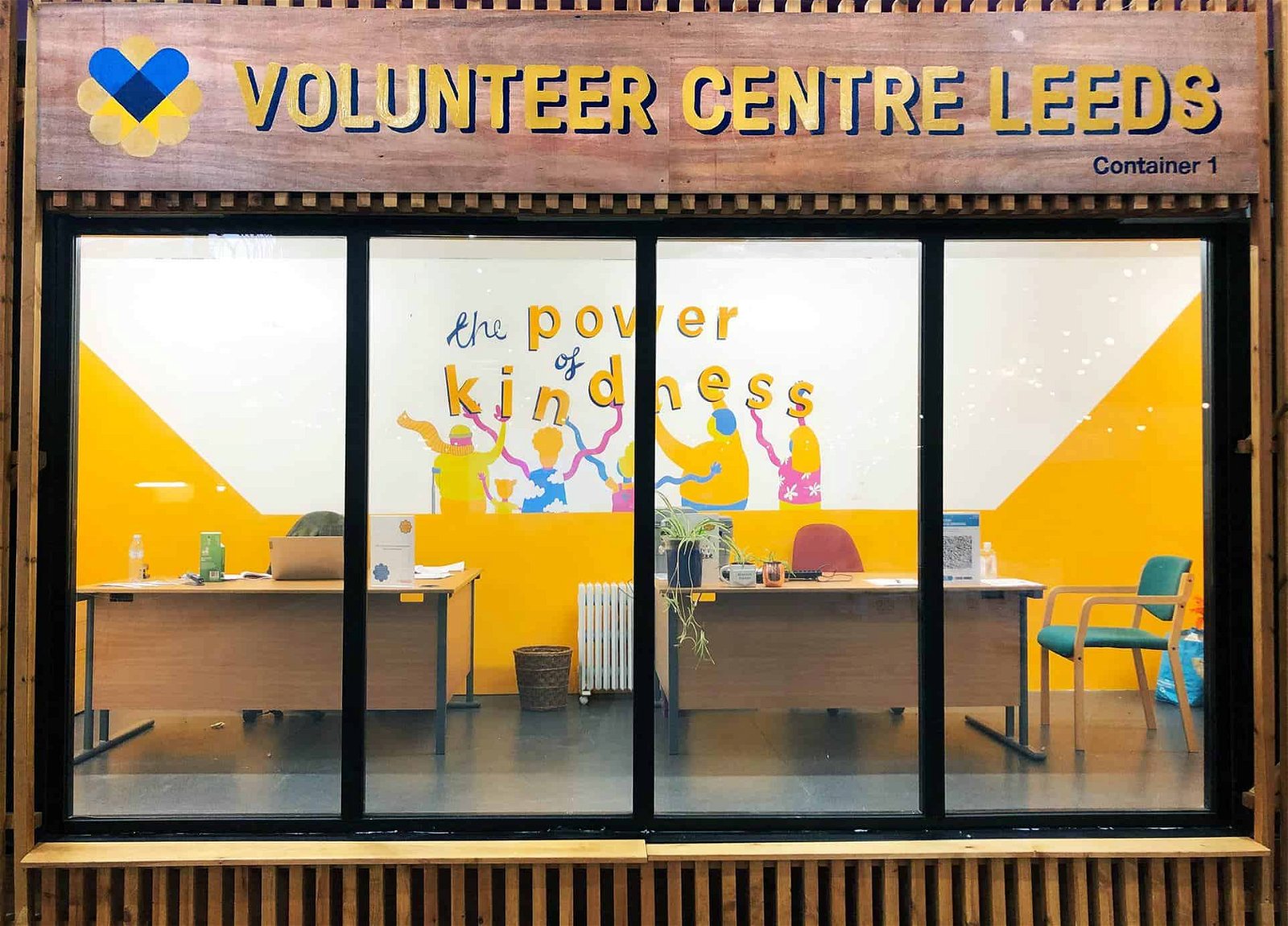 Photo of the front of the volunteer centre, there is a big glass window showing inside the centre where there are two desks and a colourful mural on the back wall which has the words the poser of kindness painted on